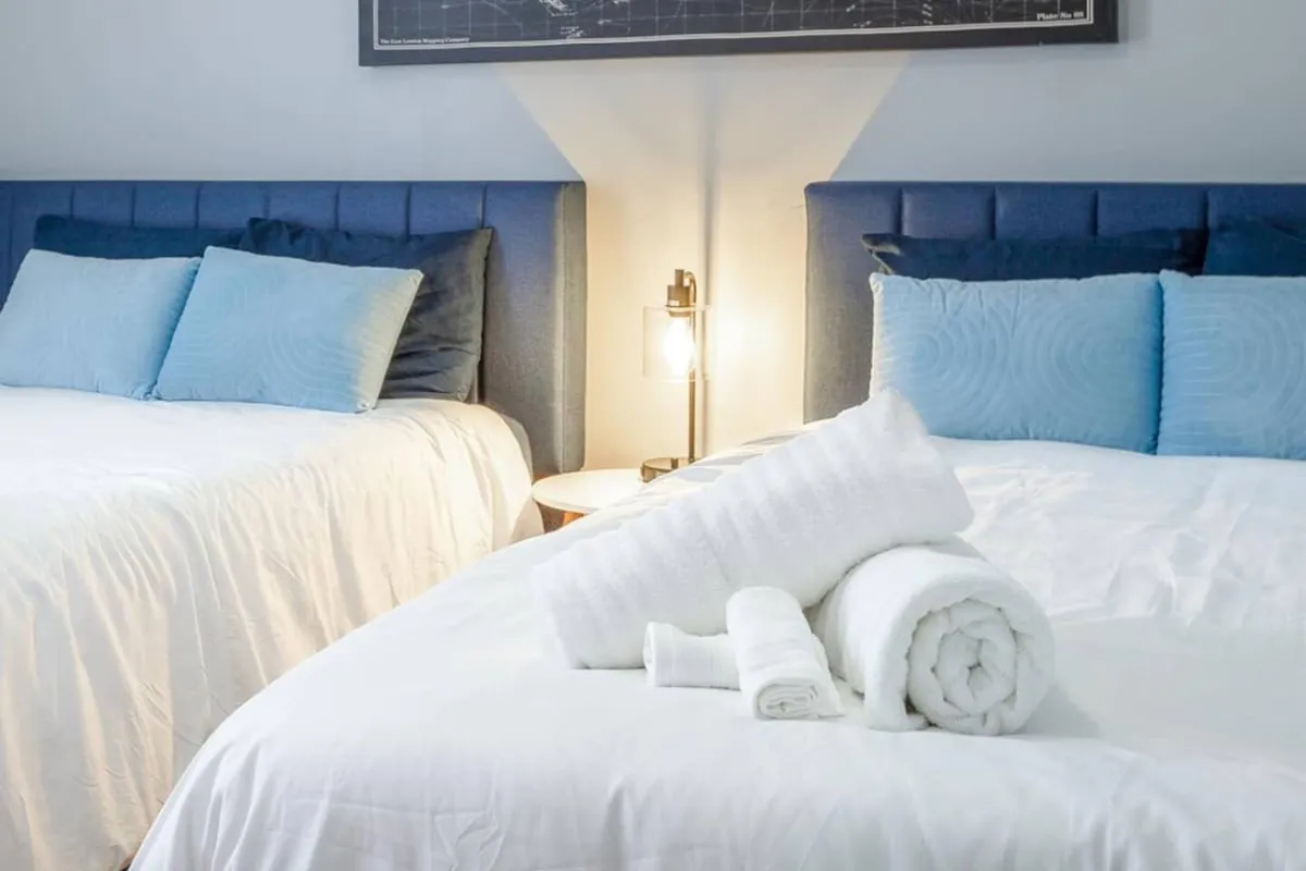 A tranquil bedroom featuring two queen beds with blue headboards, soft blue pillows, and stacked white towels. A warm lamp glows on the nightstand, with framed artwork adding a touch of personality to the serene space. The arrangement promises both comfort and flexibility for multiple guests.