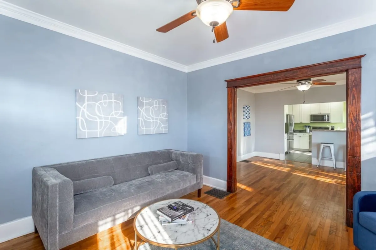 The image features a cozy living area with a grey sofa, marble coffee table, tasteful artwork on the walls, and an open kitchen area in the background. The space is bathed in natural light, creating an inviting and relaxing setting, perfect for socializing, reading, or unwinding.