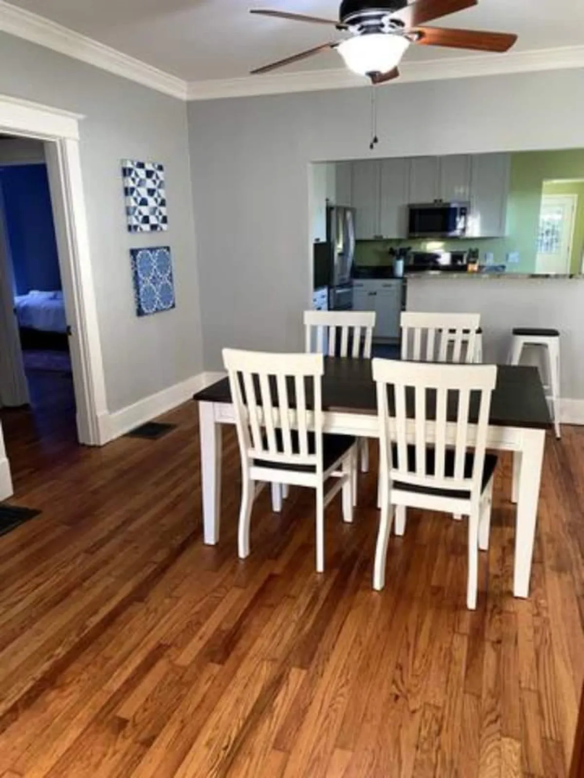 A warm and welcoming dining area is showcased, complete with a wooden dining table, elegant floor, wall art, and a clear view into the open kitchen. The overall layout feels spacious and communal, with plenty of natural light filling the space—ideal for meals, games, and conversation.