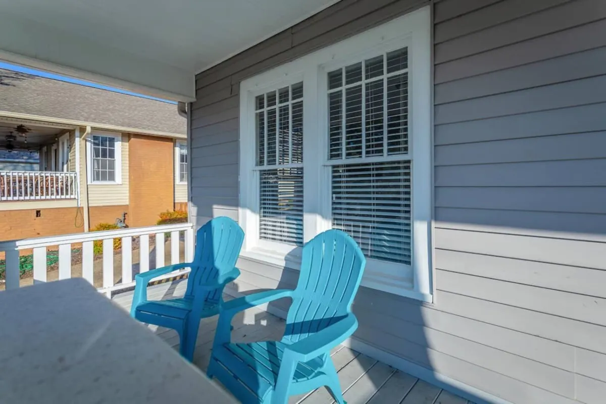 The image features two blue Adirondack chairs placed outside a window with white shutters, against the gray siding of the house. The setting is casual and relaxed, with the chairs positioned to take in sunrise views, afternoon shade, or an evening breeze, suggesting a tranquil environment ideal for unwinding after remote work or for reflective moments.