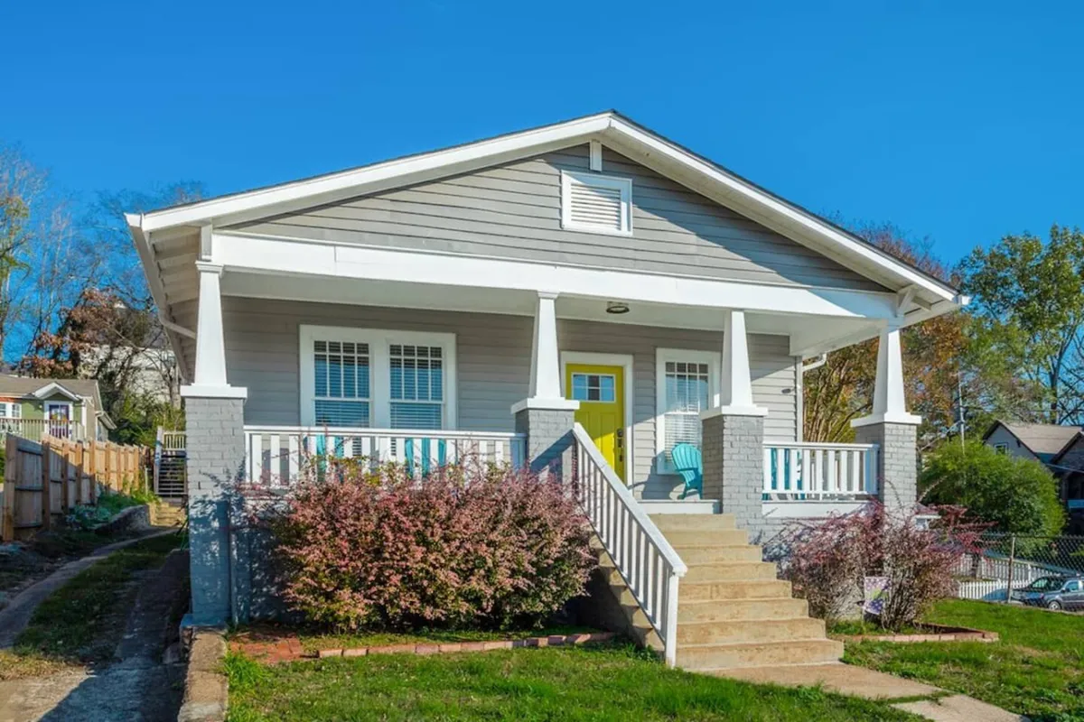 An exterior view featuring a bright yellow door framed by a landscaped garden and a spacious front porch, radiating a cheerful, welcoming vibe. The setting suggests a lively, community-oriented street just steps from downtown attractions.