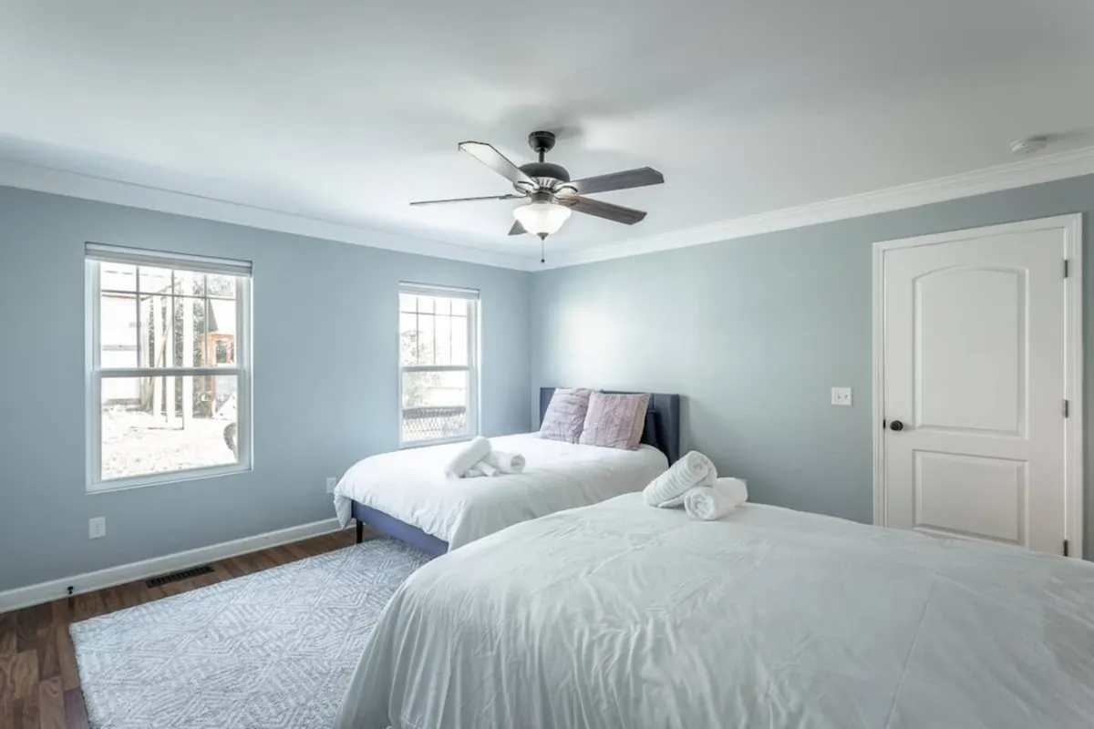 A serene bedroom with two queen beds dressed in crisp white linens, illuminated by soft morning light streaming through generous windows. A ceiling fan adds comfort, and the clean, uncluttered layout ensures privacy and restful sleep for all guests.
