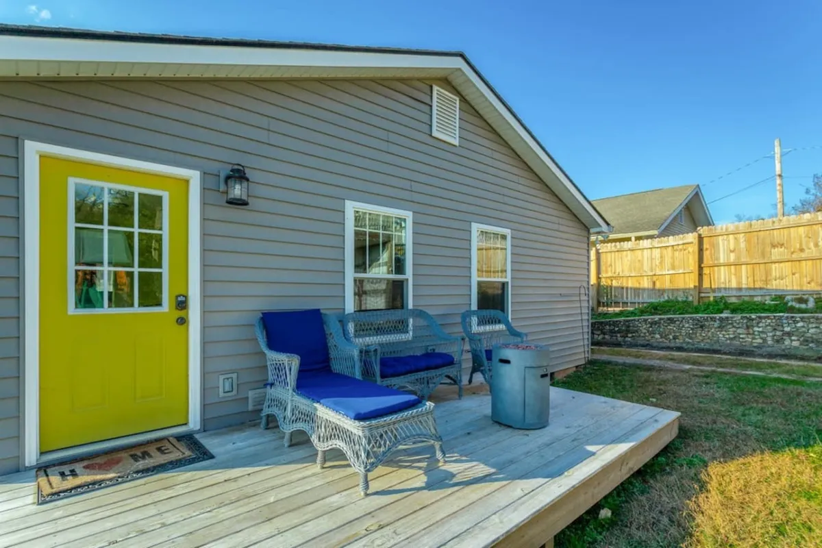 This image presents the home's inviting wooden deck with woven chairs and a bright yellow door. The deck is bathed in natural light, suggesting a peaceful spot to enjoy morning coffee or unwind at sunset. The space directly connects the indoors to the outdoors, evoking the experience of relaxing or recharging while taking in fresh air and neighborhood ambiance.
